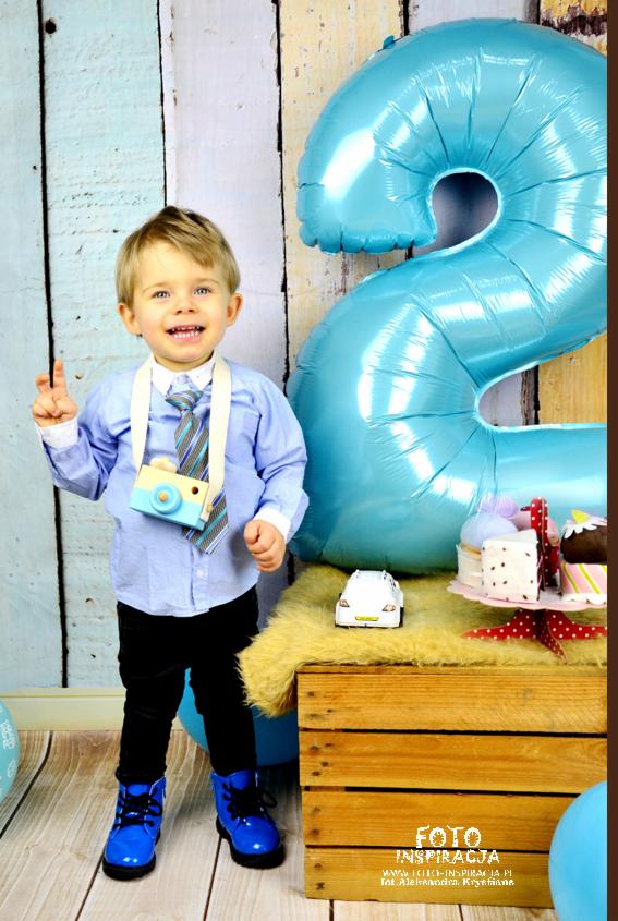 Uśmiechnięty chłopiec w błękitnej koszuli, czarnych spodniach i lakierowanych butach, z drewnianym aparatem na szyi, pozuje obok dekoracji urodzinowej z balonem w kształcie cyfry '2', drewnianej...