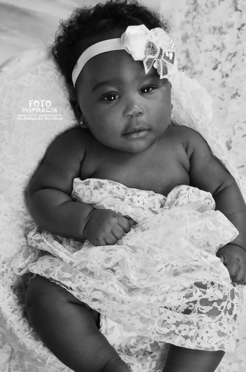 Czarno-białe zbliżenie na noworodka z ciemną karnacją, owiniętego w koronkową tkaninę i z opaską z kokardą na głowie, leżącego na wzorzystym tle.