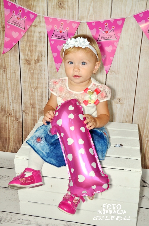 Dziewczynka z blond włosami i białą opaską siedzi na białej skrzyni, trzymając różowy balon w kształcie cyfry jeden, różowe buty, tło z drewnianych paneli i girlandy z napisem Princess.