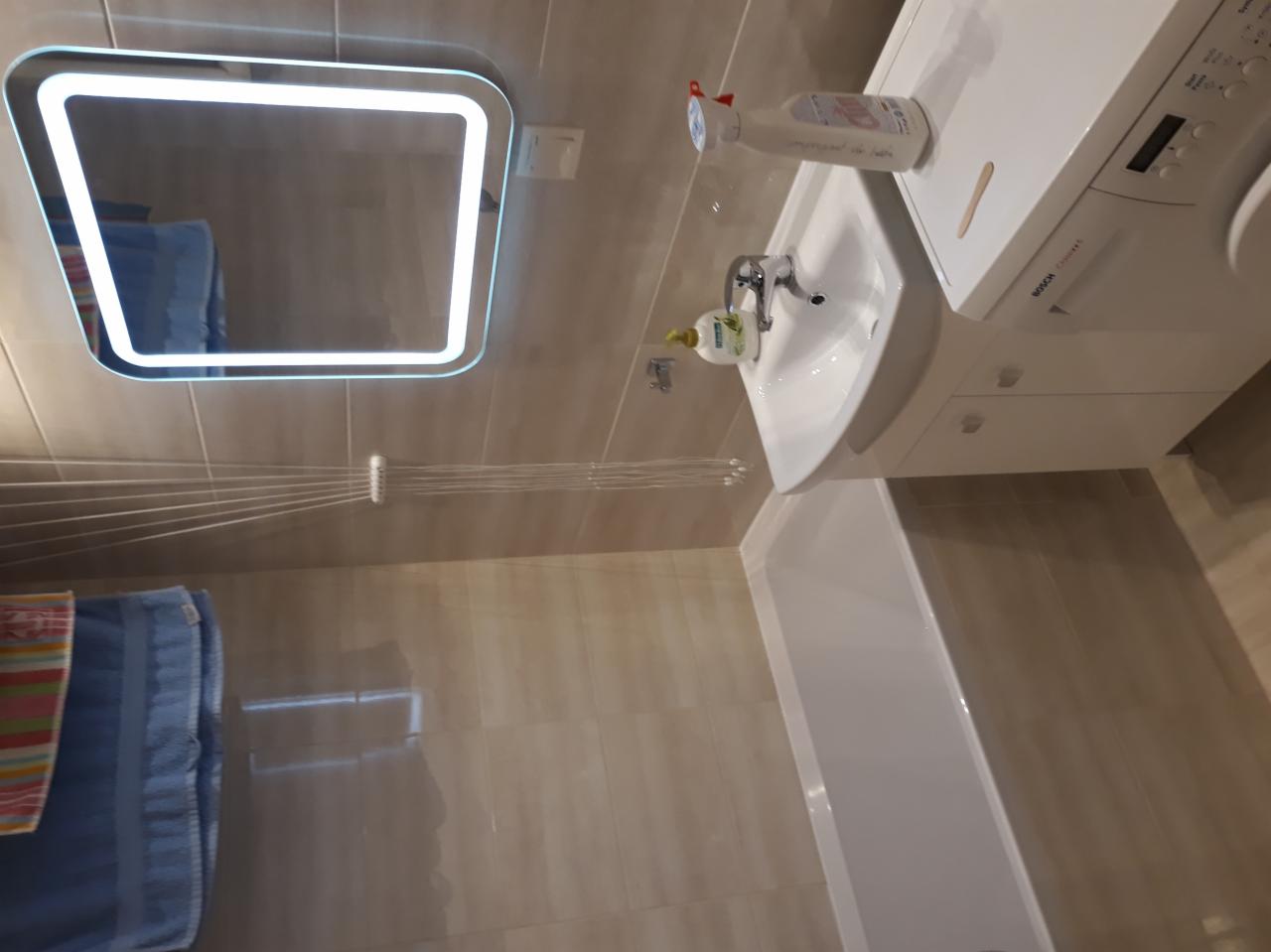 Umywalka z szafką i pralką w łazience z podświetlanym lustrem i suszarką sufitową, widoczne środki czystości.