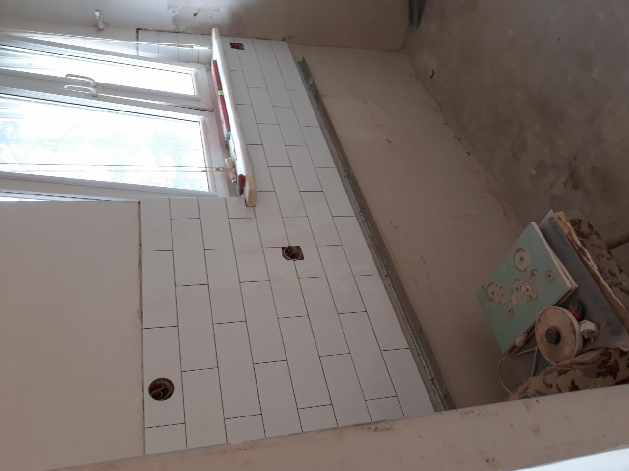 Układanie białych, podłużnych płytek w łazience, widoczny poziomica przy oknie i otwory na gniazdka elektryczne, w tle widoczna szlifierka kątowa do płytek.