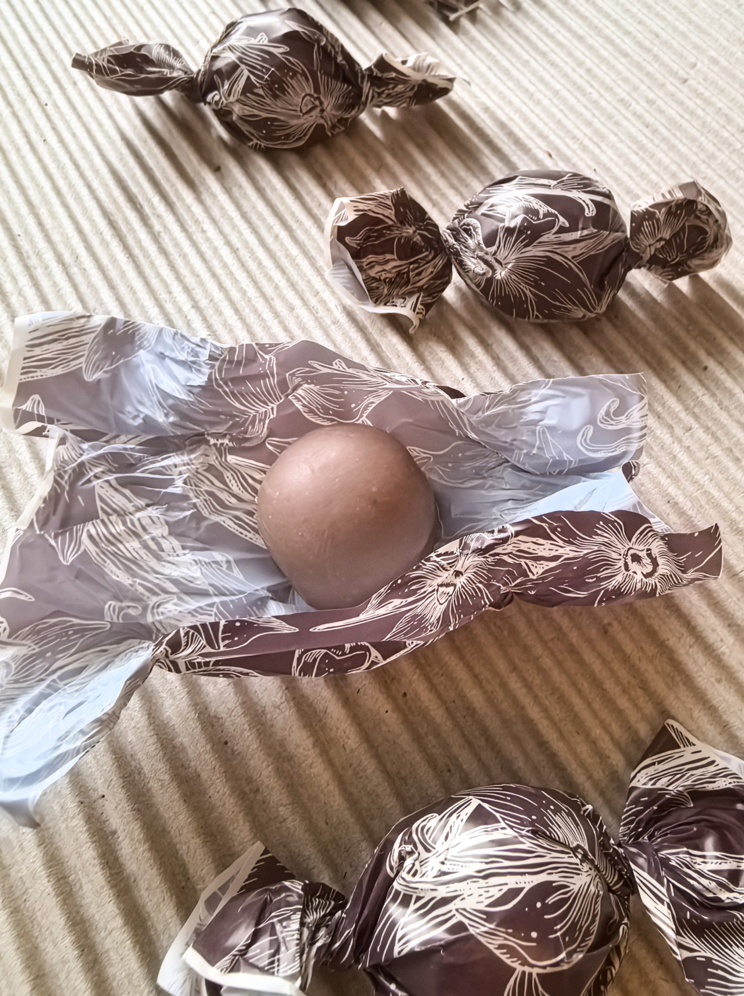 Cukierki w ozdobnych, brązowo-białych opakowaniach z motywem kwiatowym, jeden otwarty, ukazujący czekoladową kulę. Folia mineralowa Smart Paper.