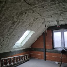 Symeria Sp. z o.o. - Poddasze po natrysku izolacji piankowej, widoczne okno dachowe, ceglana ściana, drabina i przewody. Izolacja pokrywa skośny sufit i fragment ściany.