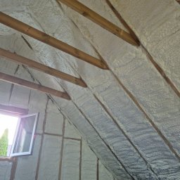 Symeria Sp. z o.o. - Poddasze ocieplone pianką, widoczne drewniane krokwie i okno. Izolacja równomiernie pokrywa powierzchnię, tworząc jednolitą warstwę. Widok z dołu na skośny dach.