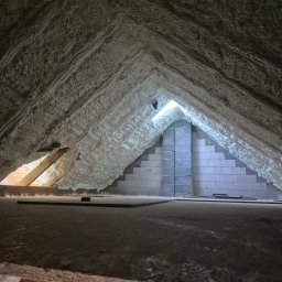 Symeria Sp. z o.o. - Poddasze po ociepleniu pianką PUR: biała, nierówna powierzchnia izolacji na skośnych ścianach i suficie, widoczne bloczki betonowe i fragment drewnianej konstrukcji.