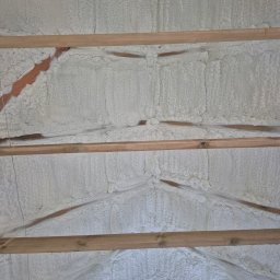 Symeria Sp. z o.o. - Ocieplenie poddasza pianką natryskową, widoczne drewniane belki konstrukcyjne. Aplikacja piany w trudno dostępnych miejscach, równomierna warstwa izolacji.