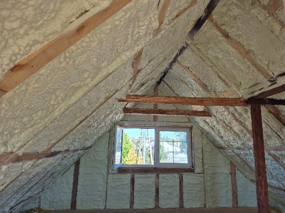 Poddasze po natrysku izolacji. Widoczne drewniane elementy konstrukcji dachu i okno. Izolacja pokrywa skośne ściany i sufit, tworząc jednolitą warstwę.
