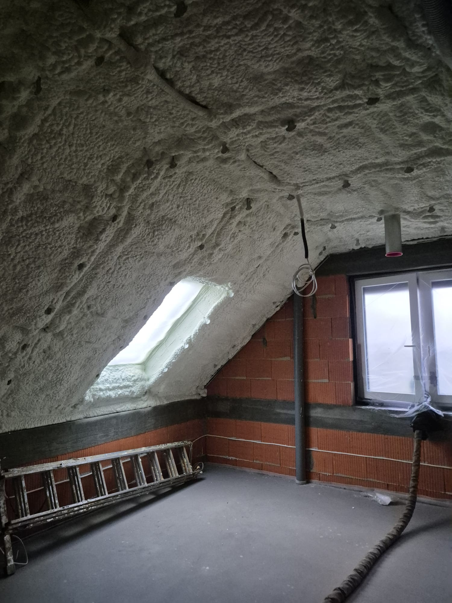 Poddasze po natrysku izolacji piankowej, widoczne okno dachowe, ceglana ściana, drabina i przewody. Izolacja pokrywa skośny sufit i fragment ściany.