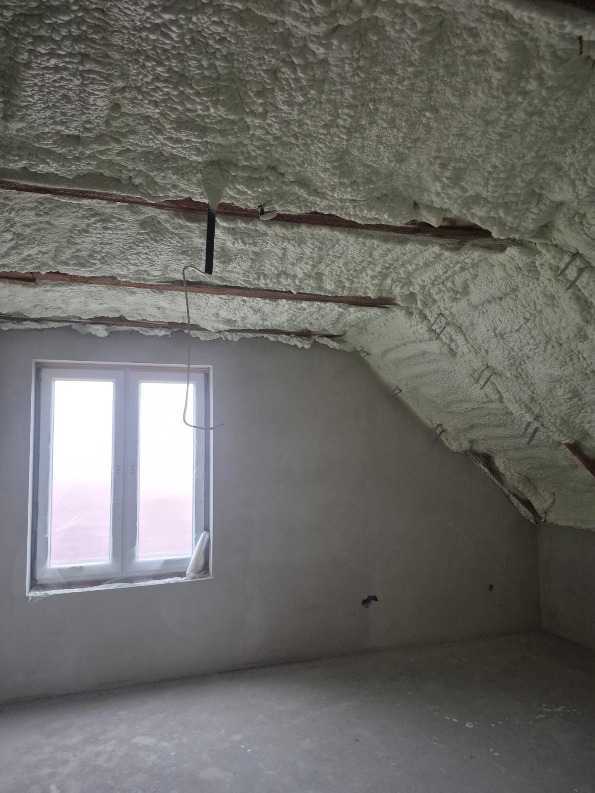 Poddasze po natrysku izolacji. Widoczne drewniane elementy konstrukcji dachu pokryte pianą, okno w szczytowej ścianie. Szara, surowa ściana i podłoga.