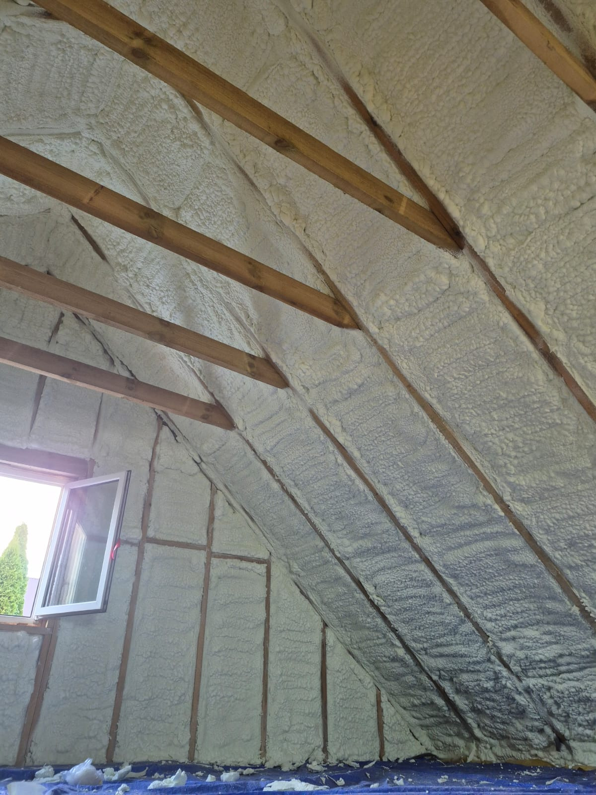 Poddasze ocieplone pianką, widoczne drewniane krokwie i okno. Izolacja równomiernie pokrywa powierzchnię, tworząc jednolitą warstwę. Widok z dołu na skośny dach.