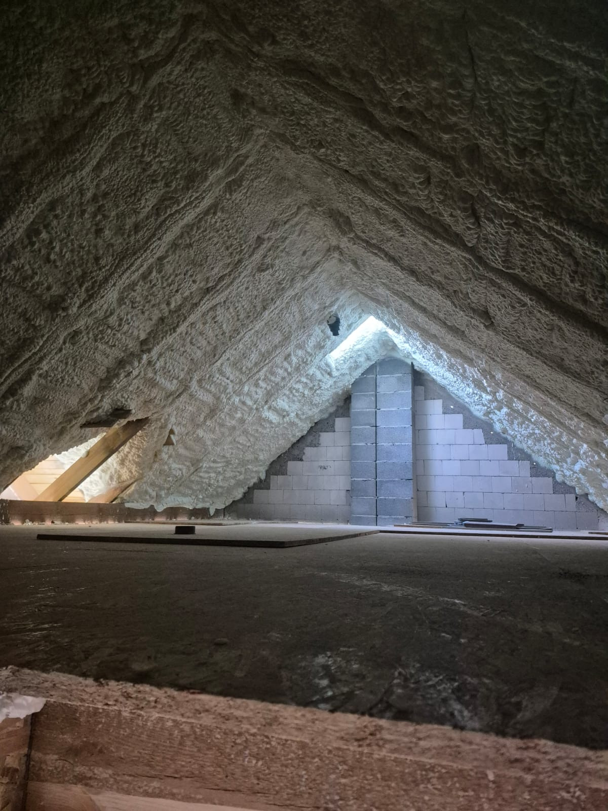 Poddasze po ociepleniu pianką PUR: biała, nierówna powierzchnia izolacji na skośnych ścianach i suficie, widoczne bloczki betonowe i fragment drewnianej konstrukcji.