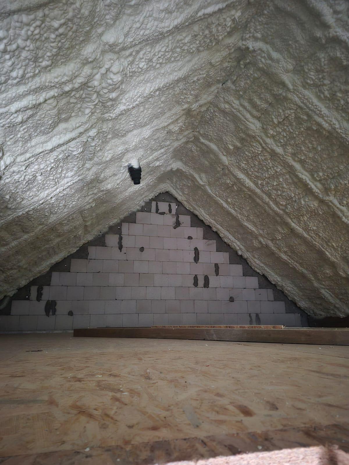 Poddasze ocieplone pianą, widok z dołu. Biała, nierówna powierzchnia piany pokrywa skośne ściany i sufit, kontrastując z szarą ścianą z pustaków i drewnianą podłogą.