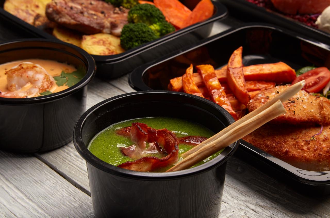 Zestaw dań w czarnych pojemnikach na wynos: zielona zupa z bekonem, zupa krem z krewetkami, mięso z ziemniakami i brokułami, panierowana ryba ze słodkimi ziemniakami i pomidorem, ułożone na szarym...