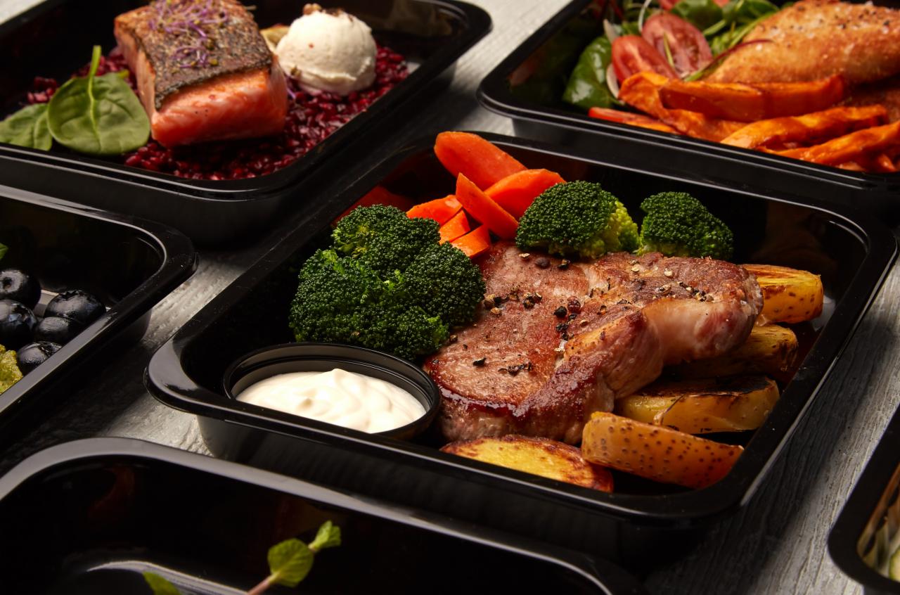 Zestaw posiłków w czarnych pojemnikach: stek z brokułami i marchewką, łosoś na czerwonym ryżu z kremowym musem, sałatka ze słodkimi ziemniakami i borówki.