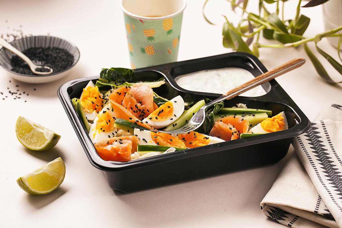 Dietetyczny lunchbox z łososiem, jajkiem na twardo, ogórkiem i sosem tzatziki, posypany czarnym sezamem, podany z limonką i napojem ananasowym w papierowym kubku.