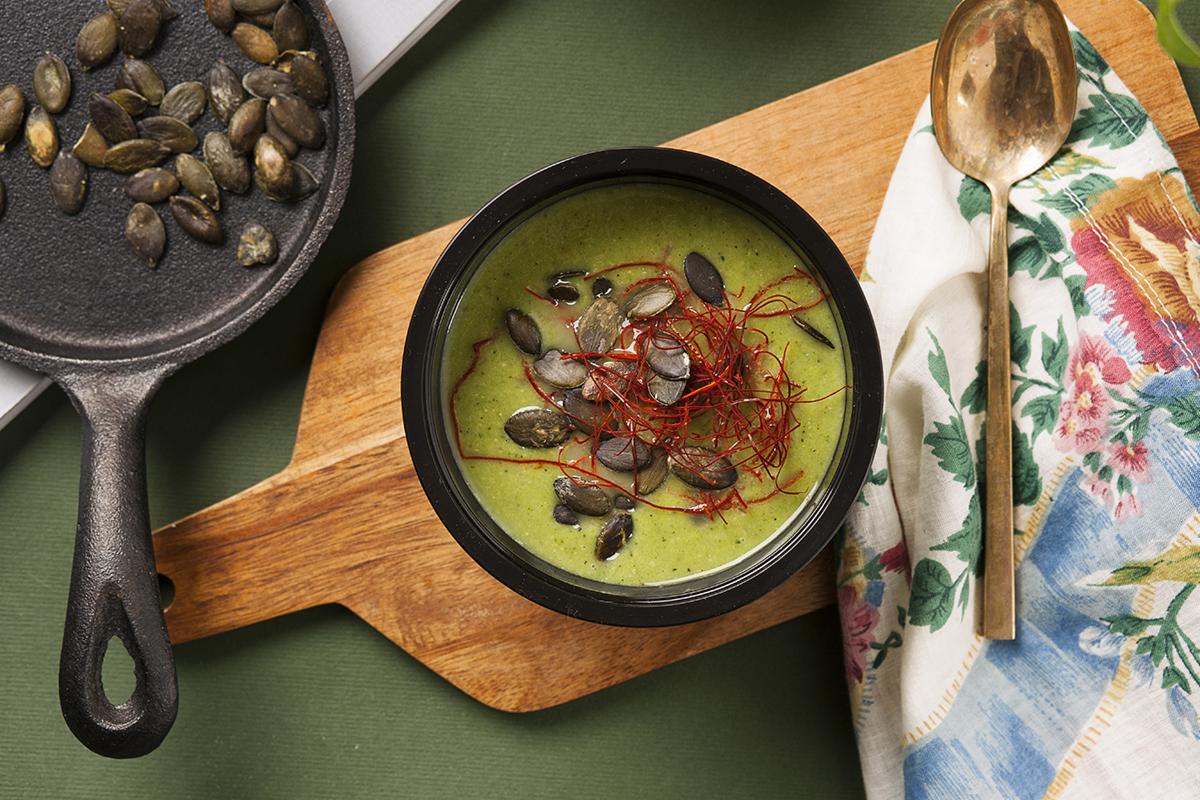 Zielony krem zupy w czarnej misce, posypany pestkami dyni i nitkami chili, na drewnianej desce obok patelni z pestkami dyni i złotej łyżki na kwiecistej serwetce.