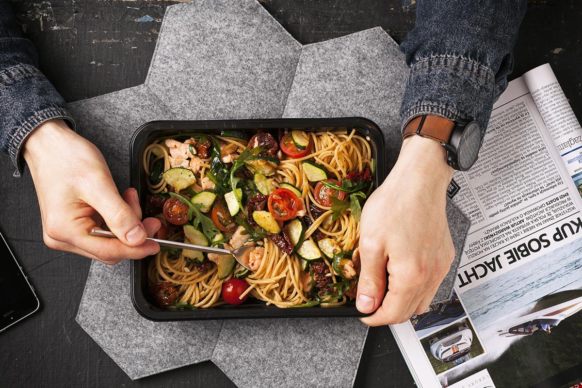 Zbliżenie na pudełko z makaronem spaghetti z warzywami i łososiem, trzymane przez osobę w dżinsowej koszuli, obok gazety z artykułem o jachtach i smartfona na szarym podkładzie.