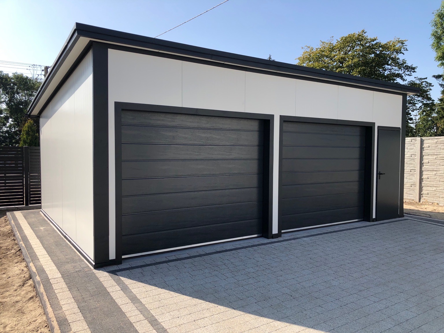 Nowoczesny garaż dwustanowiskowy z płyt warstwowych w kolorze białym z czarnymi bramami segmentowymi i drzwiami bocznymi, wykończony kostką brukową.