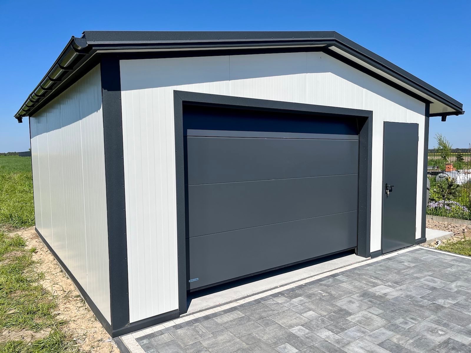 Gotowy, biało-szary garaż blaszany z bramą segmentową i drzwiami bocznymi, zrealizowany w Sulejówku, na tle zielonego pola i błękitnego nieba.