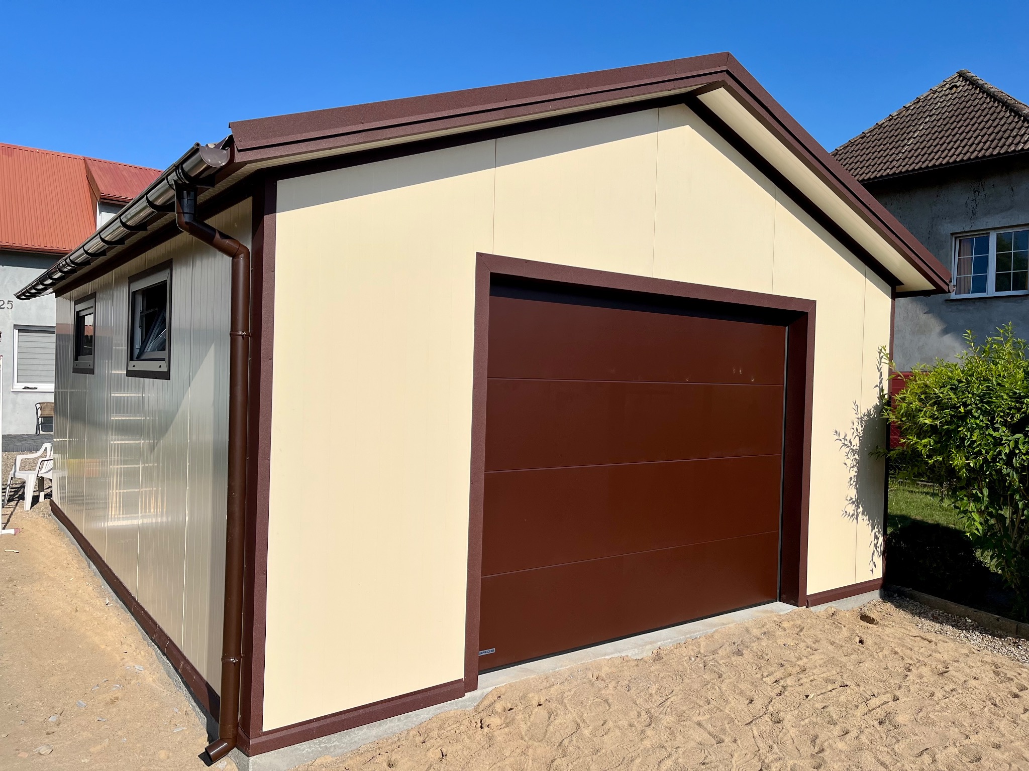 Gotowy garaż z płyt warstwowych w kolorze beżowym z brązowymi elementami wykończeniowymi, widok z zewnątrz na tle błękitnego nieba.