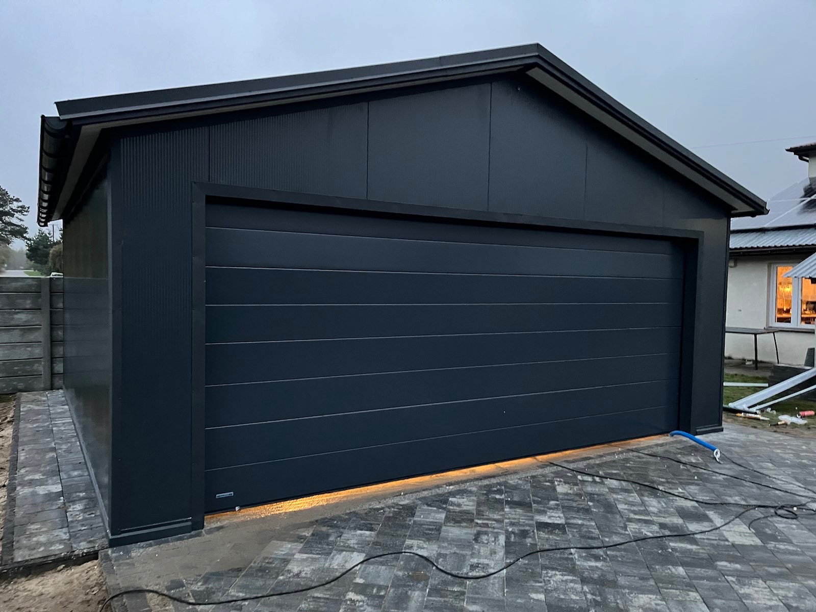 Nowoczesny garaż z ciemnoszarą segmentową bramą, wykończony płytami warstwowymi w tym samym kolorze, oświetlenie LED u dołu bramy, kostka brukowa przed garażem, pochmurne niebo.