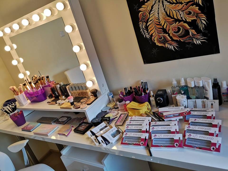 Stanowisko wizażysty z lustrem otoczonym żarówkami, pędzlami do makijażu w fioletowych pojemnikach, paletami cieni, kosmetykami Lancome i dużą ilością sztucznych rzęs marki Intervion na białym blacie.