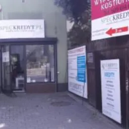 Fasada budynku z szyldem SPECKREDYT.PL, widoczna z chodnika, z przystankiem autobusowym po lewej stronie i bujną zielenią po obu stronach.