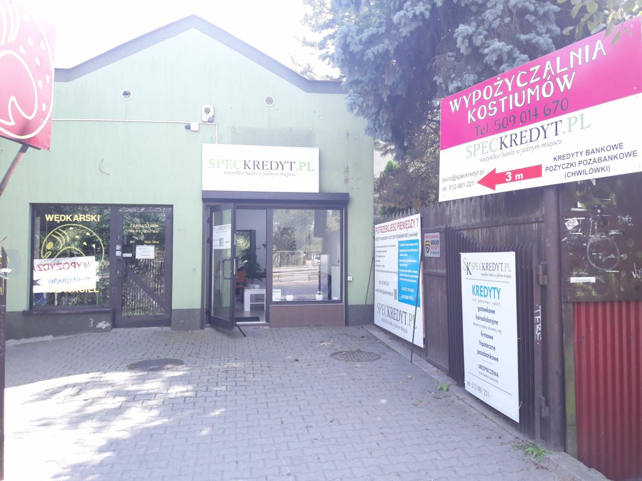 Fasada budynku z szyldem SPECKREDYT.PL, obok reklama wypożyczalni kostiumów i oferta kredytów gotówkowych na tablicach informacyjnych przy chodniku.