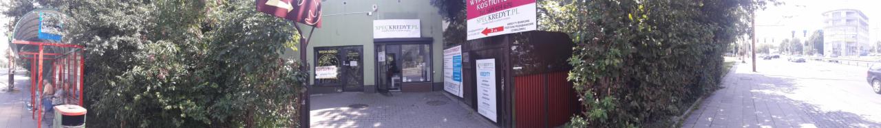 Fasada budynku z szyldem SPECKREDYT.PL, widoczna z chodnika, z przystankiem autobusowym po lewej stronie i bujną zielenią po obu stronach.