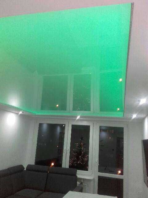 Zielony, błyszczący sufit napinany z oświetleniem LED w nowoczesnym salonie, z widokiem na świątecznie oświetloną choinkę za oknem.