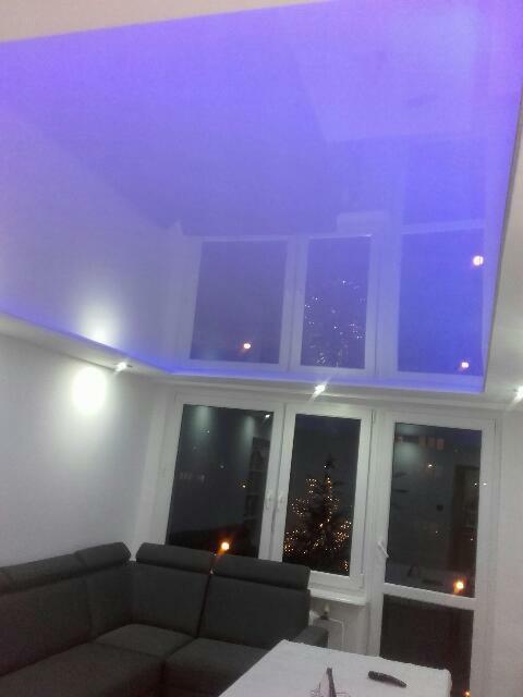 Lśniący, fioletowy sufit napinany z oświetleniem LED w nowoczesnym salonie z widokiem na miasto nocą przez okna.