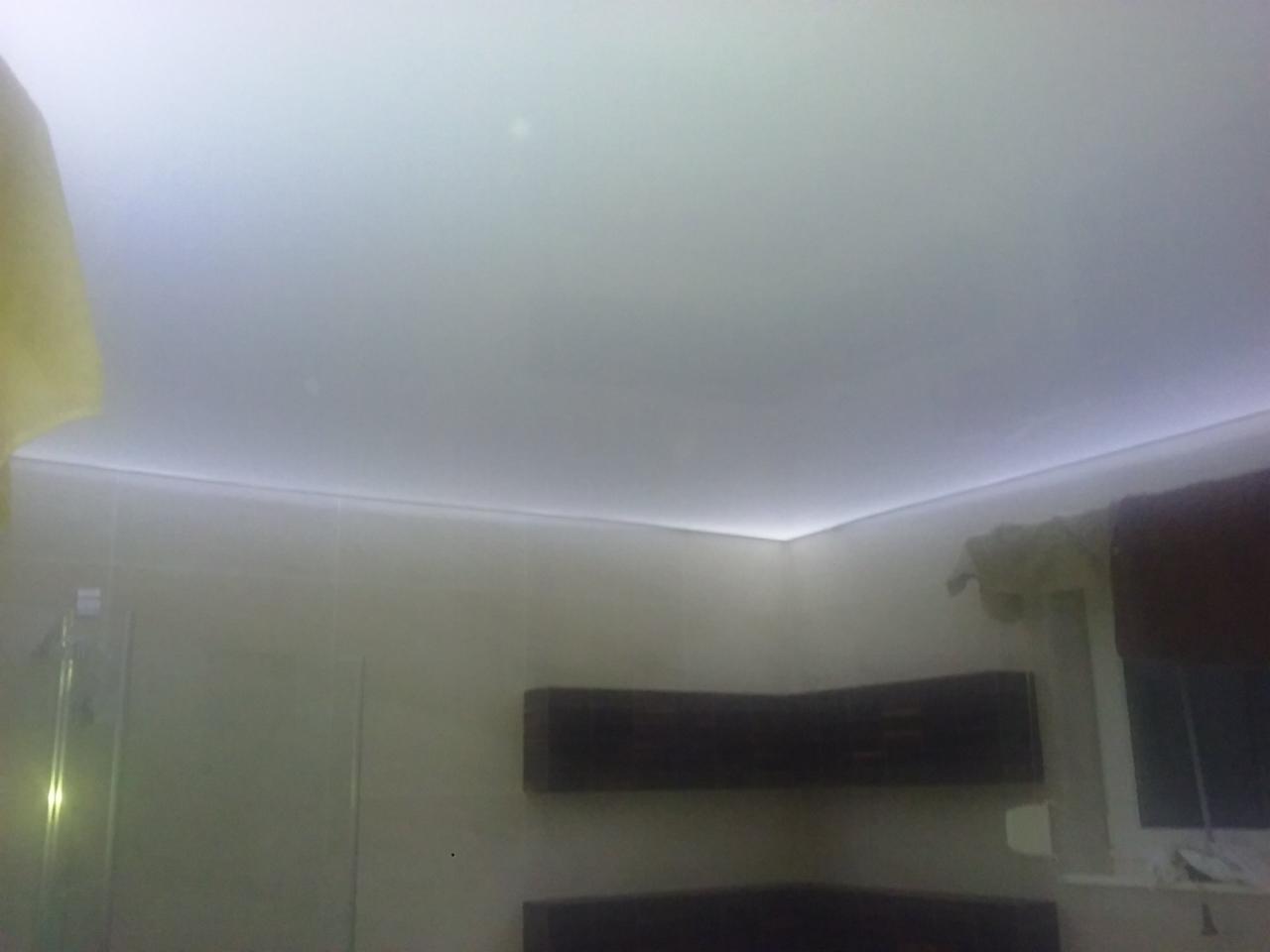 Biały sufit napinany z subtelnym oświetleniem LED po obwodzie w łazience, widoczna część ściany z szafkami i okno z zasłoną.