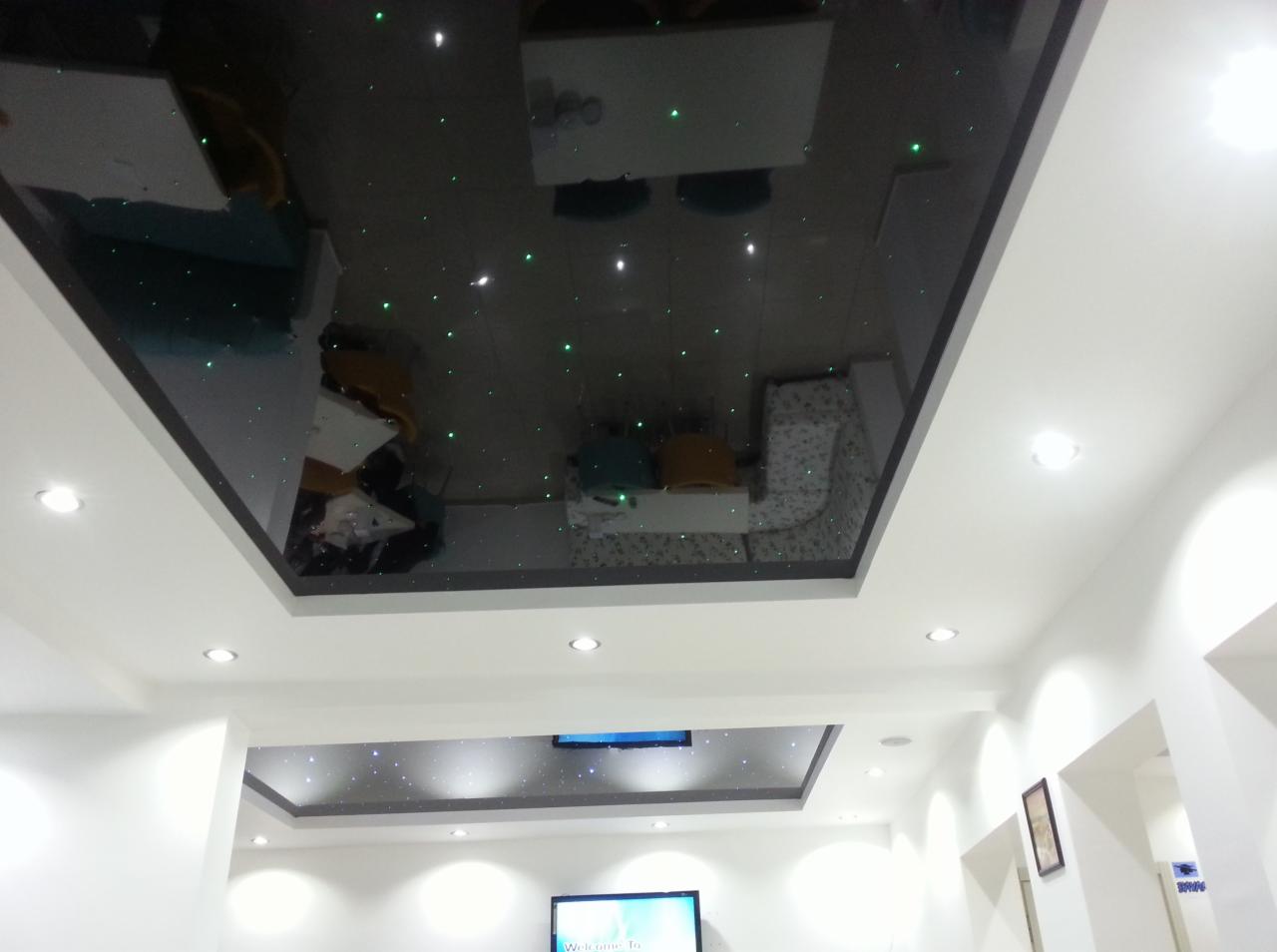 Nowoczesny salon z dwupoziomowym sufitem podwieszanym, górny poziom to czarny, błyszczący sufit napinany z zielonymi diodami imitującymi gwiazdy, dolny poziom biały z oświetleniem punktowym.