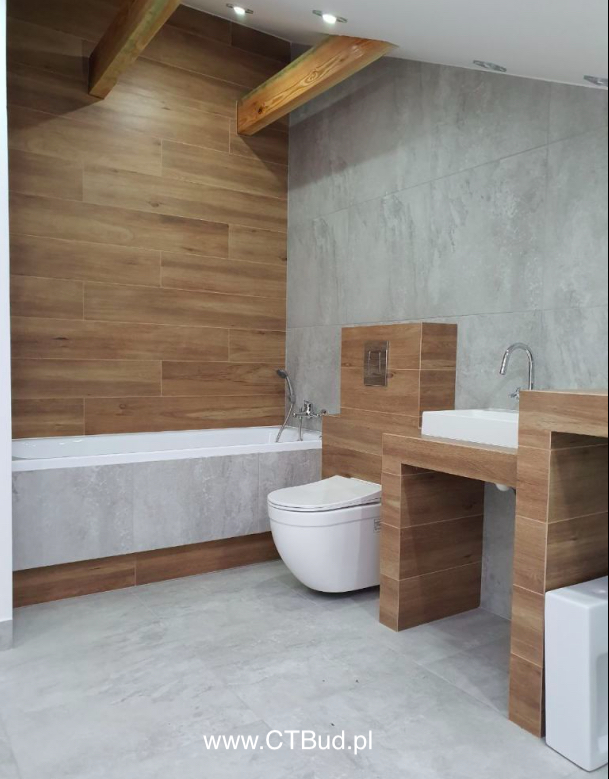 Nowoczesna łazienka z drewnopodobnymi płytkami na ścianach, drewnianymi belkami dekoracyjnymi pod sufitem, białą armaturą i szarą podłogą z płytek.