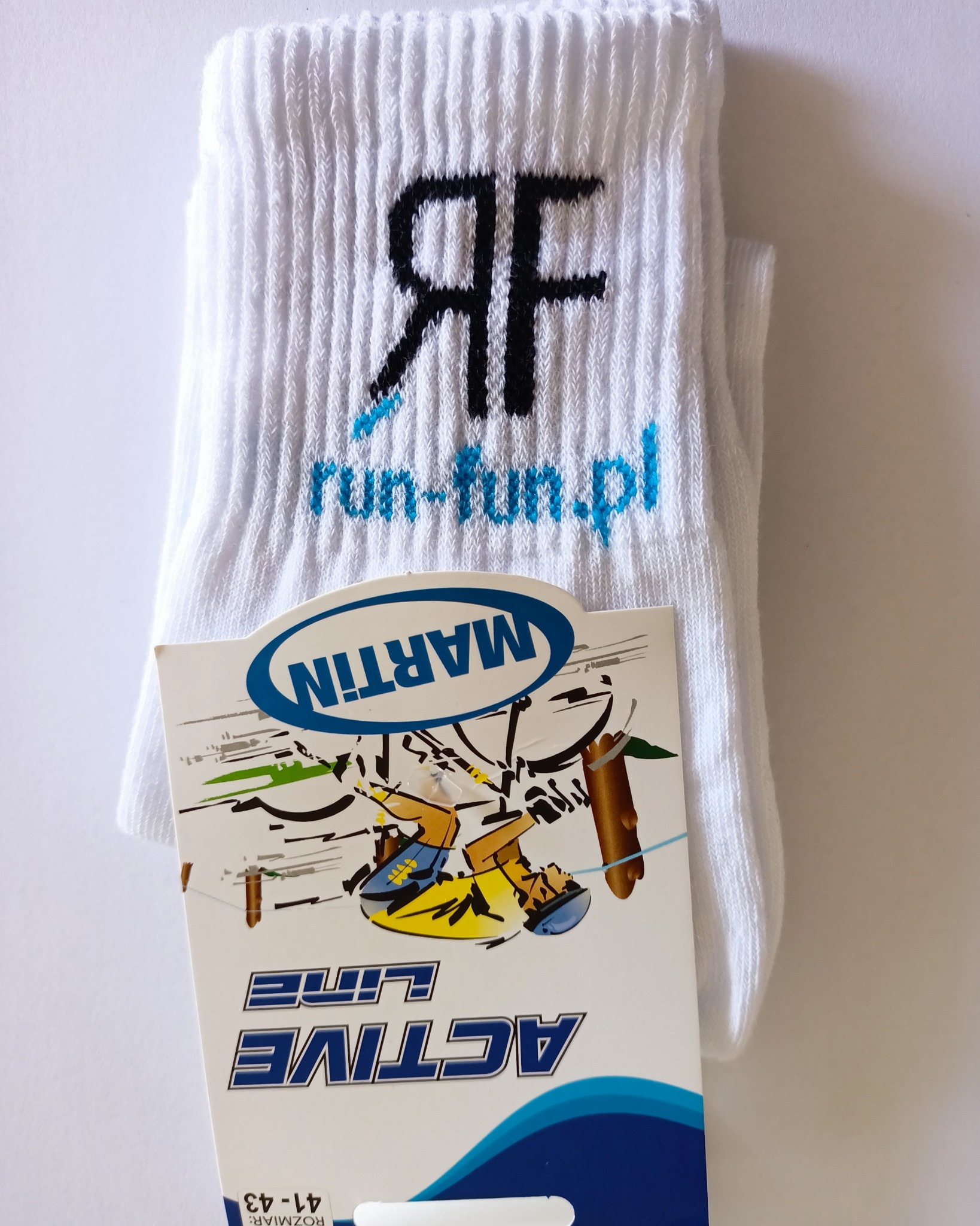 Białe, prążkowane skarpetki z czarnym logo 'RF' i niebieskim napisem 'run-fun.pl', leżące obok kartonowej etykiety z rysunkiem kolarza i napisem 'MARTIN ACTIVE LINE'.