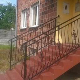 Paweł Kowalczyk - Zewnętrzne schody wejściowe do domu z cegły, zabezpieczone ozdobną, ciemną balustradą z kutego metalu z motywem kwiatowym. W tle widoczny betonowy płot.