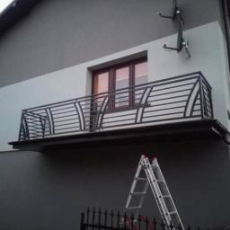 Paweł Kowalczyk - Nowoczesny balkon z kutą metalową balustradą z poziomymi prętami, widoczna drabina oparta o ścianę budynku w trakcie montażu.