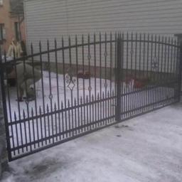 Paweł Kowalczyk - Czarna, metalowa brama wjazdowa z kutymi zdobieniami, osadzona na kamiennych słupkach, widoczna osoba pracująca w tle.