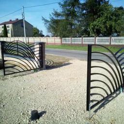 Paweł Kowalczyk - Otwarta brama wjazdowa z kutego metalu z dekoracyjnymi łukami, osadzona na kamiennych słupkach, widok na żwirowy podjazd i ogrodzenie panelowe w tle.