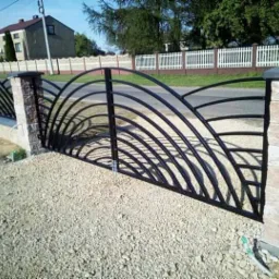 Czarna brama z łukowatymi prętami, osadzona między słupkami z jasnej cegły, prowadząca na żwirowy podjazd, w tle białe ogrodzenie i dom.