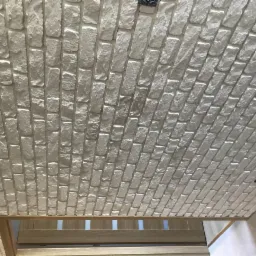 Wykończony sufit z dekoracyjnej cegły w odcieniach bieli, z widocznymi punktami oświetleniowymi i fragmentem drewnianej ramy drzwiowej w dolnej części kadru.