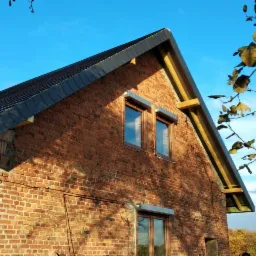 Ceglany budynek z częściowo ukończonym dachem z ciemnej blachy, widoczne elementy konstrukcyjne i okna z roletami zewnętrznymi, w tle błękitne niebo i drzewa.