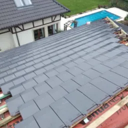 Montaż szarej dachówki na dachu domu z widokiem na basen i ogród w tle, widoczne elementy więźby dachowej i membrana dachowa z napisem OVERLAPPING LINE.