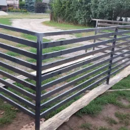 Surowa, metalowa, pozioma poręcz w trakcie montażu w Blochy, oparta na deskach, z widocznym samochodem i budynkiem gospodarczym w tle.