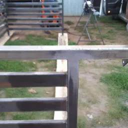 Spawana metalowa konstrukcja ogrodzenia w trakcie montażu z widocznymi elementami mocującymi, spawarką w tle i samochodem z otwartymi drzwiami.