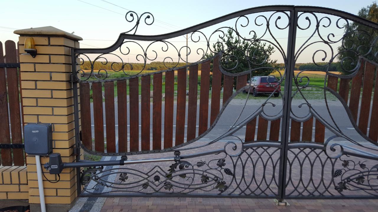 Kuta brama dwuskrzydłowa z drewnianym wypełnieniem, automatyką i ceglanym słupkiem, widok z zewnątrz na podjazd i polne tło.