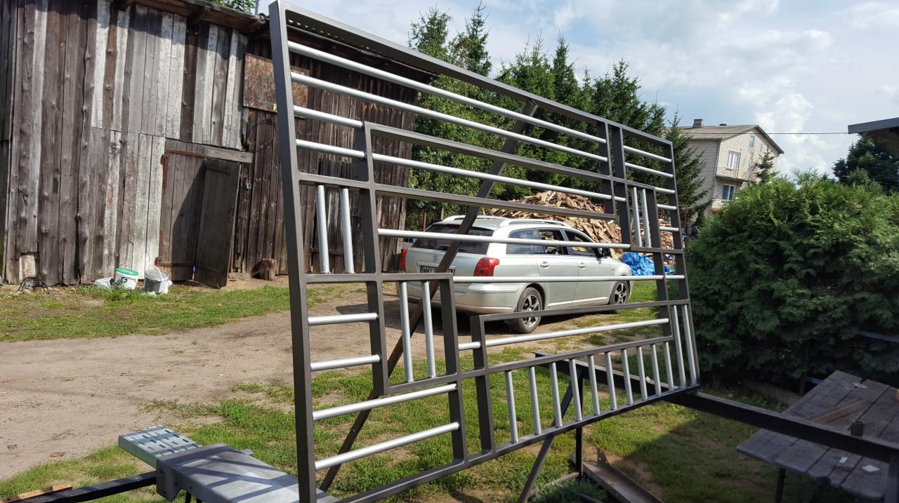 Szkielet metalowej konstrukcji z poprzecznymi prętami na tle podwórka z drewnianym budynkiem gospodarczym i zaparkowanym srebrnym kombi.