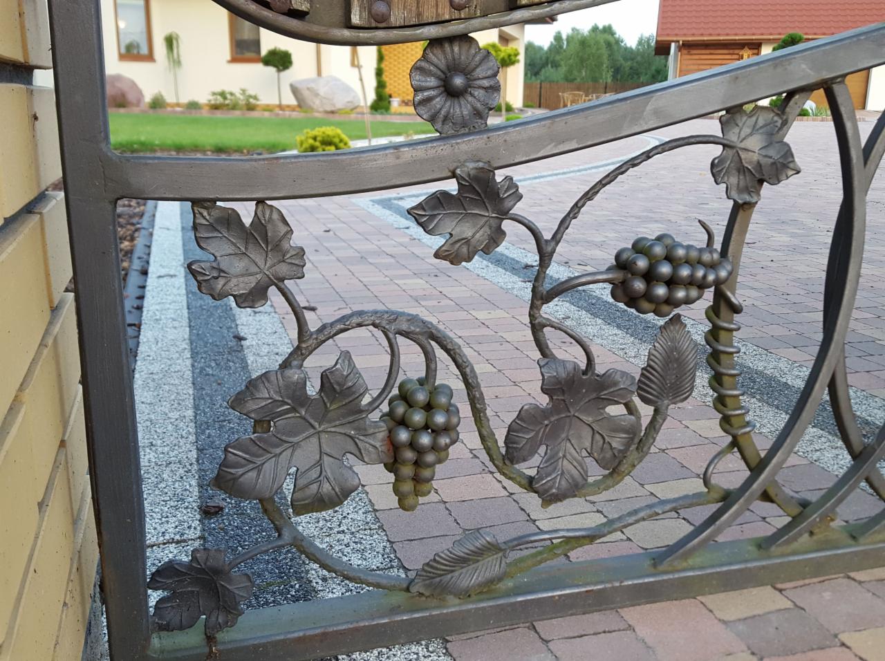Metalowa brama z kutymi elementami winorośli i gron, na tle kostki brukowej i domu z ogrodem.