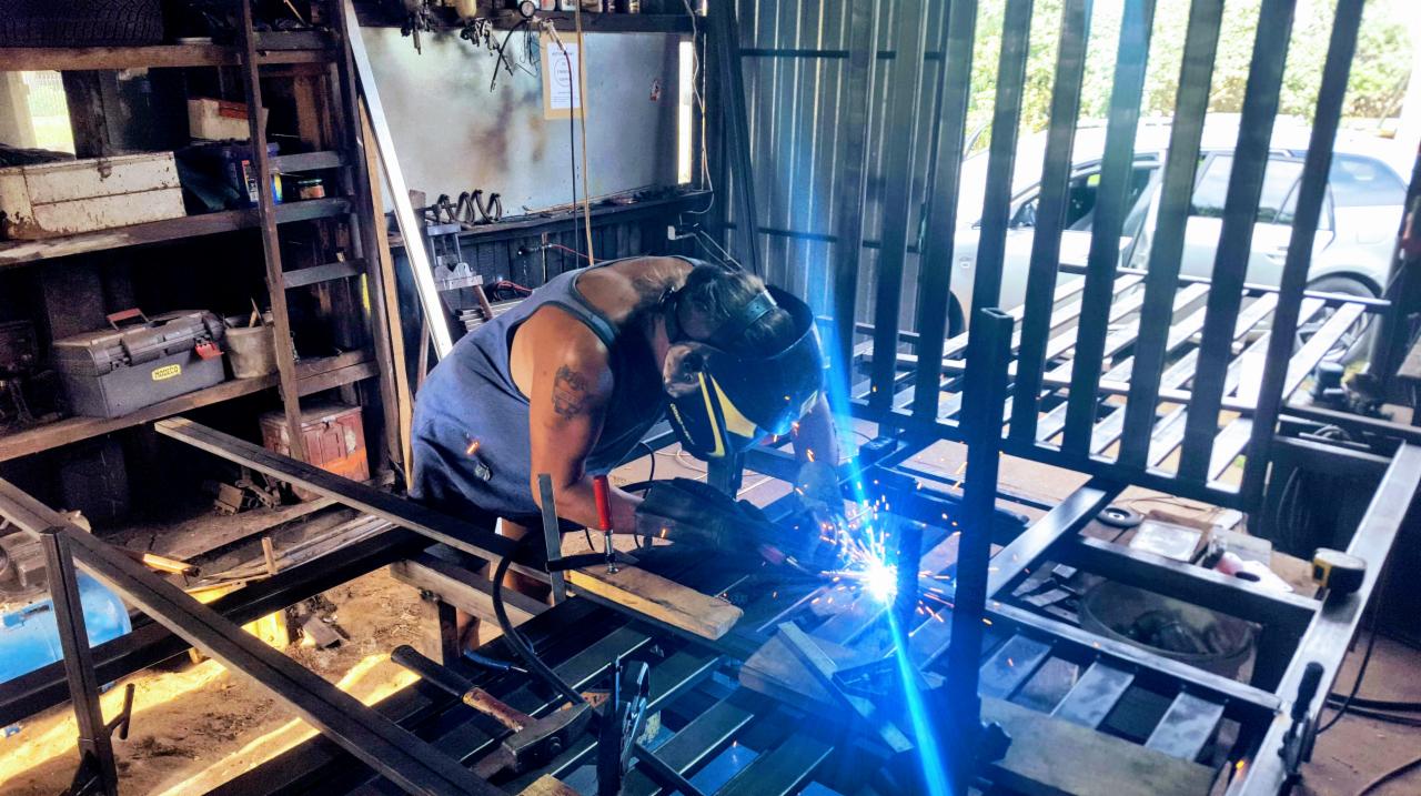 Spawacz w masce spawalniczej łączy metalowe elementy konstrukcji bramy w warsztacie, widoczne iskry i narzędzia.