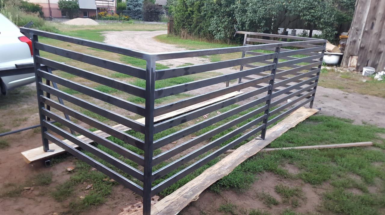 Surowa, metalowa, pozioma poręcz w trakcie montażu w Blochy, oparta na deskach, z widocznym samochodem i budynkiem gospodarczym w tle.